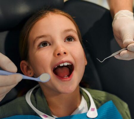 Young girl receiving dental check-up, sitting in dentist chair with open mouth, while dentist holds dental tools, representing pediatric dentistry services.