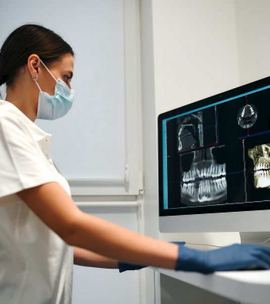 A young woman dentist in a medical mask examines an x-ray image on a computer in a dental office with modern equipment. Caries treatment. Dentistry and dental care.